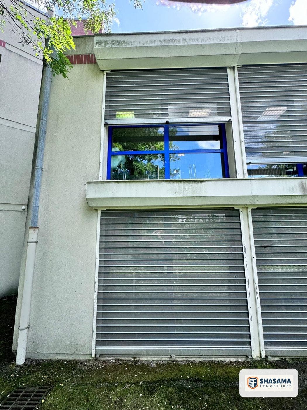 Deux rideaux métalliques fermés en façade d’immeuble, avec une fenêtre à encadrement bleu