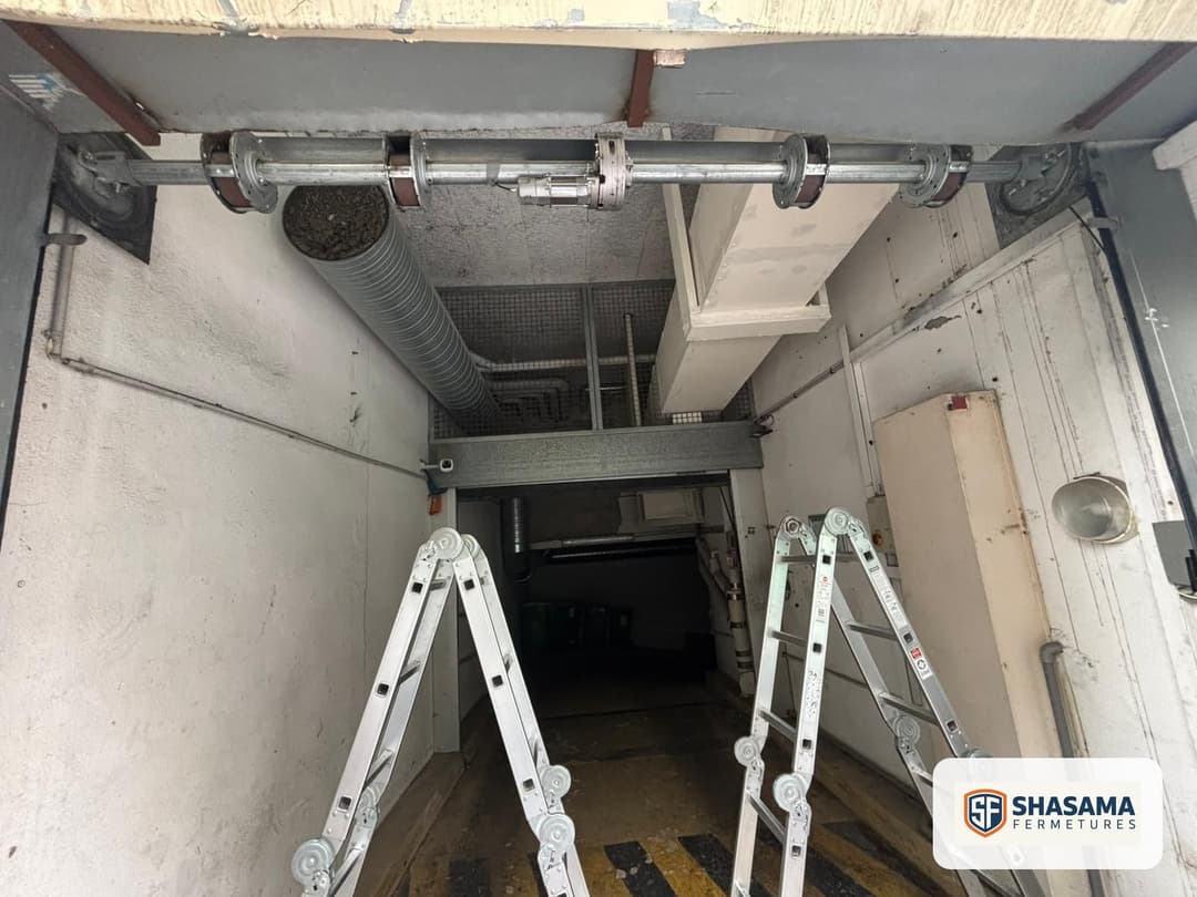 Coffre plafond d'un rideau métallique ouvert : tube d'enroulement, paliers, motoréducteur et gaine technique, échelles télescopiques au sol.