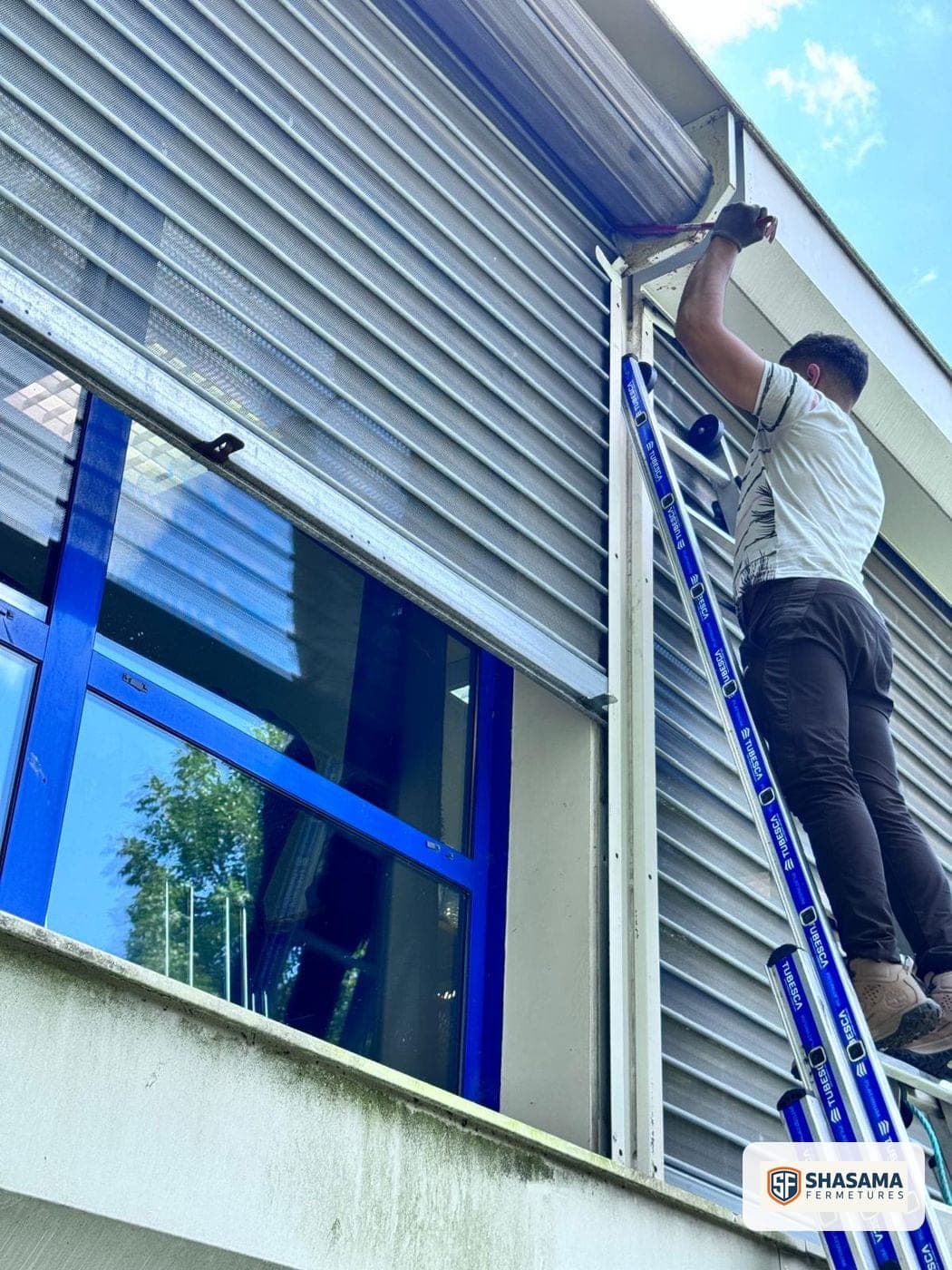Technicien Shasama Fermetures en intervention sur une échelle, ajustant le haut d’un rideau métallique le long d’une façade vitrée