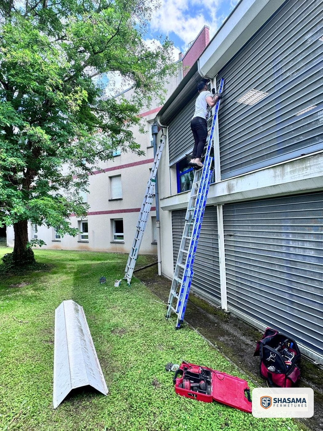 Technicien Shasama Fermetures sur une échelle, réglant le haut d’un rideau métallique le long d’une façade