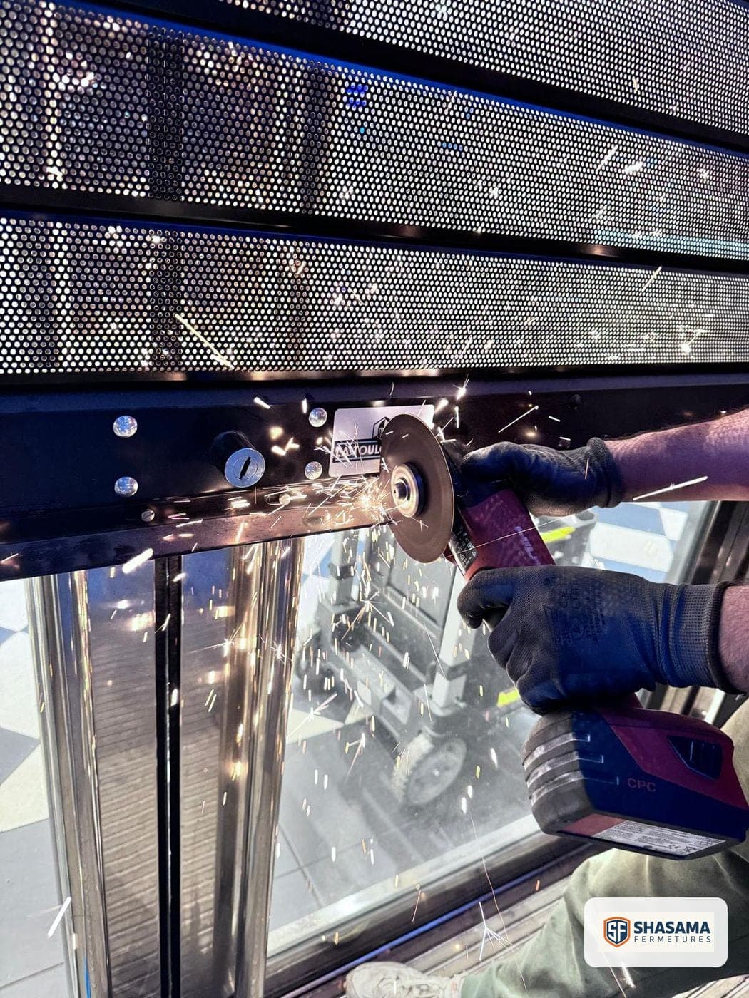 Gros plan sur les mains d’un technicien Shasama Fermetures meulant un axe de verrouillage de rideau métallique, étincelles visibles